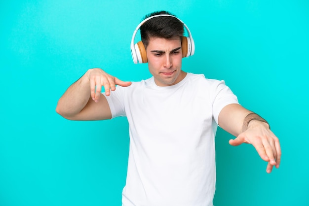 Giovane uomo caucasico isolato su sfondo blu ascoltando musica e ballando