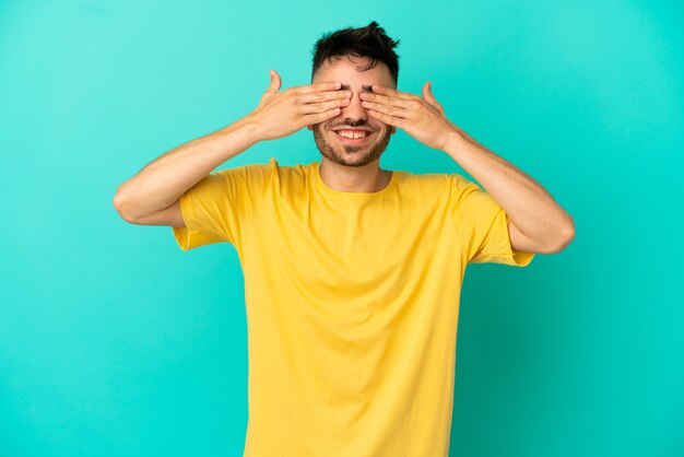 Giovane uomo caucasico isolato su sfondo blu che copre gli occhi con le mani e sorride