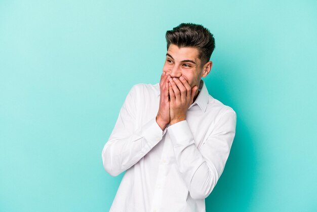 Giovane uomo caucasico isolato su sfondo blu che ride di qualcosa che copre la bocca con le mani