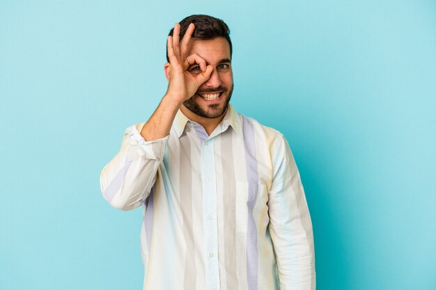 Giovane uomo caucasico isolato su sfondo blu eccitato mantenendo il gesto giusto sull'occhio.