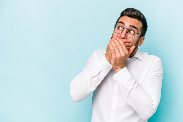 Giovane uomo caucasico isolato su sfondo blu premuroso guardando uno spazio di copia che copre la bocca con la mano