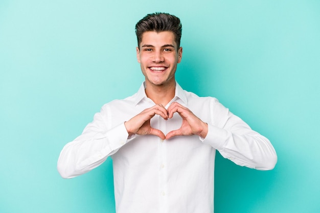 Giovane uomo caucasico isolato su sfondo blu sorridente e che mostra una forma di cuore con le mani.