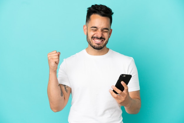 Giovane uomo caucasico isolato su sfondo blu utilizzando il telefono cellulare e facendo gesto di vittoria