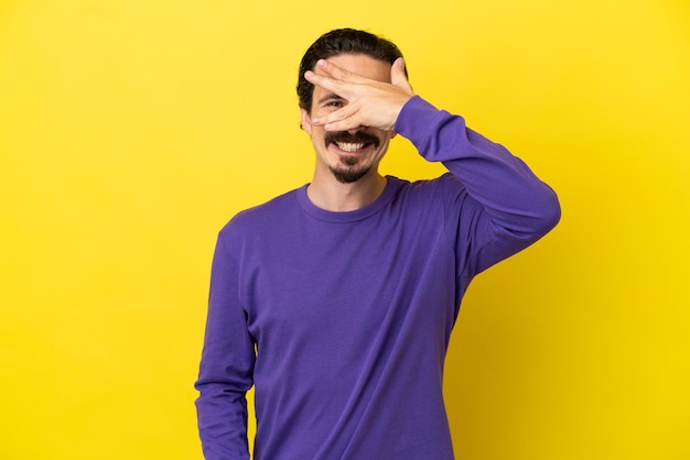 Giovane uomo caucasico isolato su sfondo giallo che copre gli occhi con le mani e sorride