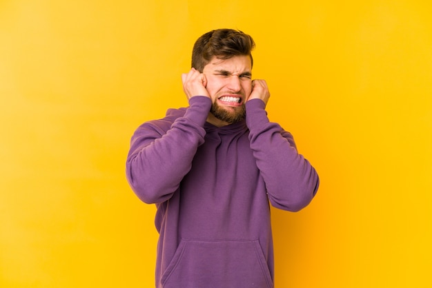 Giovane uomo caucasico isolato su sfondo giallo che copre le orecchie con le mani.