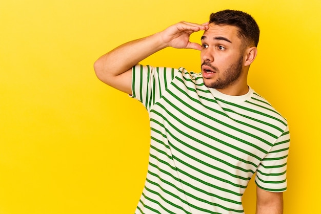 Giovane uomo caucasico isolato su sfondo giallo che guarda lontano tenendo la mano sulla fronte.