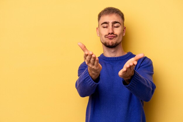 Giovane uomo caucasico isolato su sfondo giallo che piega le labbra e tiene i palmi delle mani per inviare un bacio d'aria.