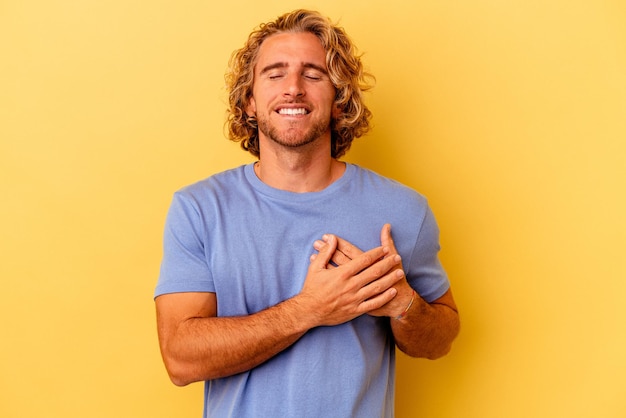 Giovane uomo caucasico isolato su sfondo giallo che ride tenendo le mani sul cuore, concetto di felicità.