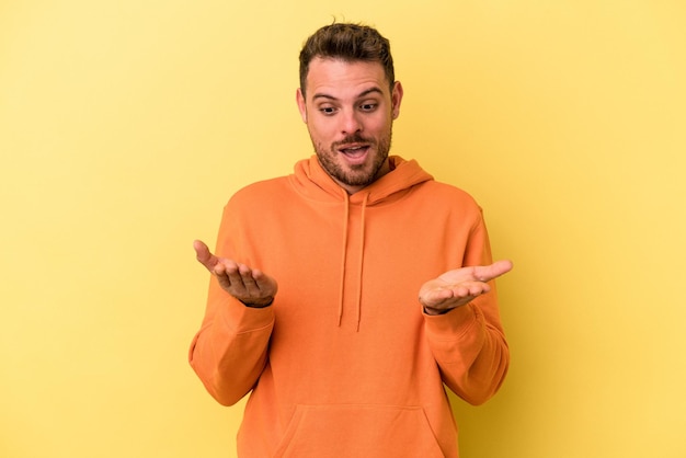 Giovane uomo caucasico isolato su sfondo giallo che tiene qualcosa con le palme, offrendo alla macchina fotografica.