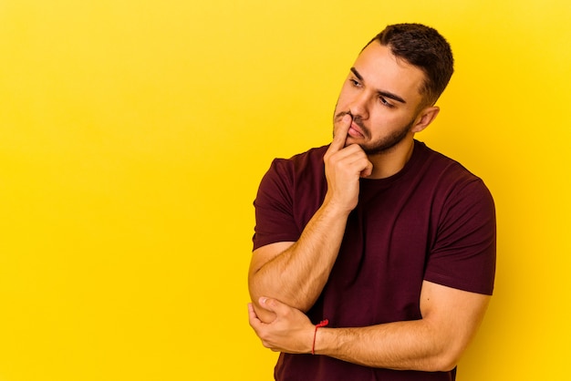 Giovane uomo caucasico isolato su sfondo giallo guardando lateralmente con espressione dubbiosa e scettica.