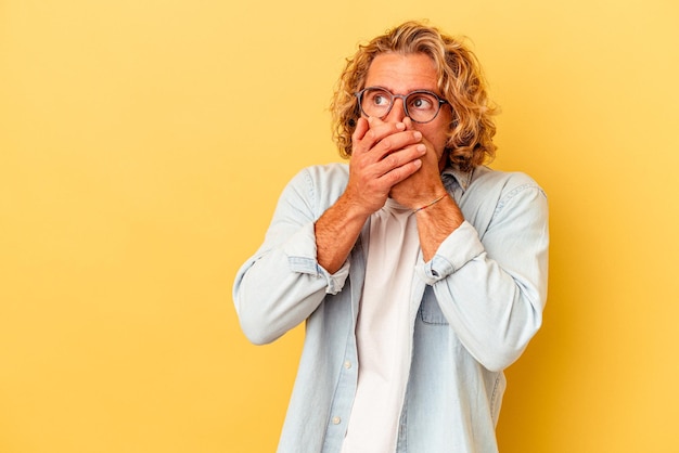 Giovane uomo caucasico isolato su sfondo giallo premuroso guardando uno spazio di copia che copre la bocca con la mano.