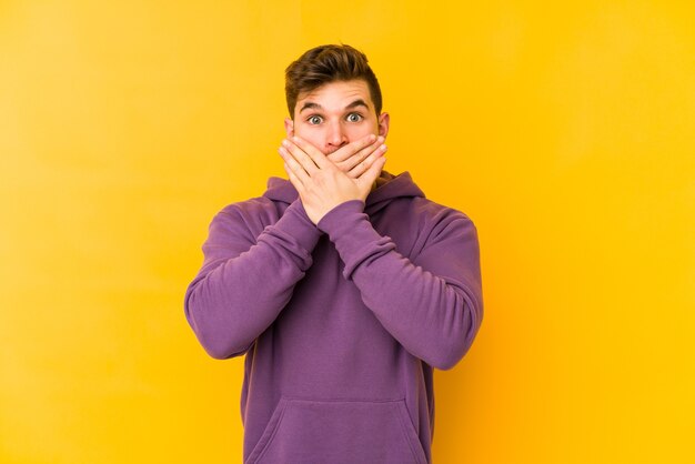 Giovane uomo caucasico isolato su sfondo giallo scioccato che copre la bocca con le mani.