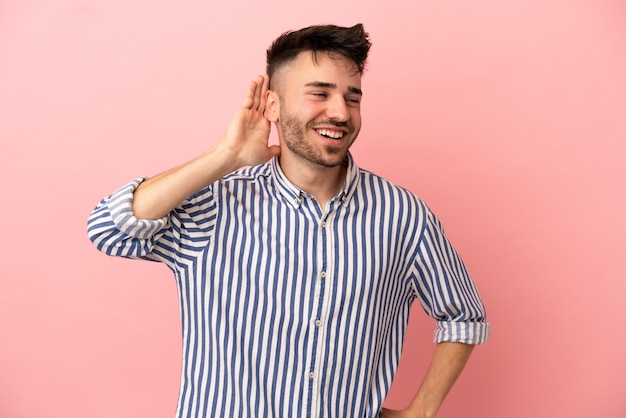 Giovane uomo caucasico isolato su sfondo rosa ascoltando qualcosa mettendo la mano sull'orecchio