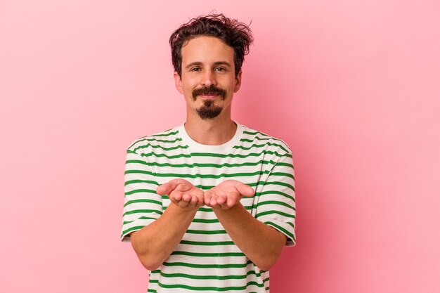 Giovane uomo caucasico isolato su sfondo rosa che tiene qualcosa con le palme, offrendo alla macchina fotografica.