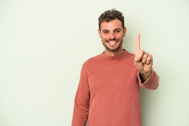 Giovane uomo caucasico isolato su sfondo verde che mostra il numero uno con il dito.