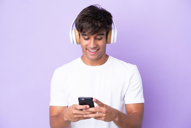 Giovane uomo caucasico isolato su sfondo viola ascoltando musica e guardando al cellulare