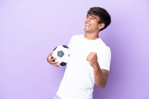 Giovane uomo caucasico isolato su sfondo viola con pallone da calcio che celebra una vittoria