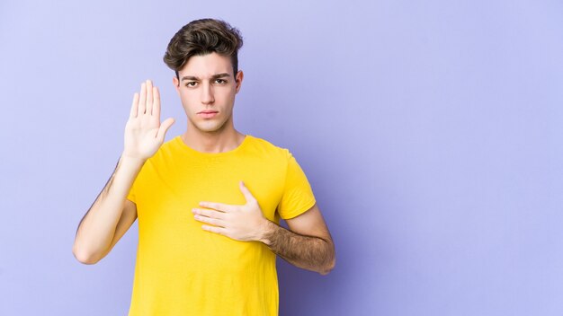 Giovane uomo caucasico isolato su viola prestando giuramento, mettendo la mano sul petto.