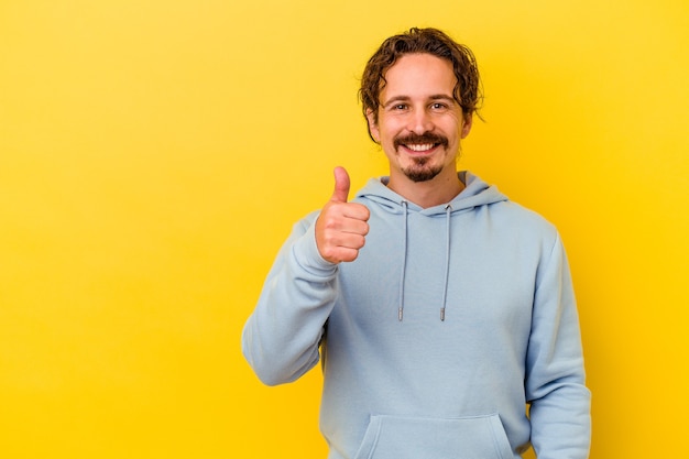 Giovane uomo caucasico isolato sul muro giallo sorridendo e alzando il pollice