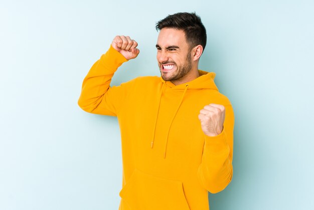 Giovane uomo caucasico isolato sulla parete blu alzando il pugno dopo una vittoria, concetto vincitore.