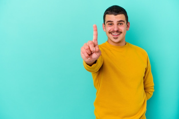 Giovane uomo caucasico isolato sulla parete blu che mostra il numero uno con il dito.