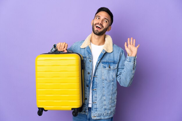 Giovane uomo caucasico isolato sulla parete blu in vacanza con la valigia di viaggio e salutando