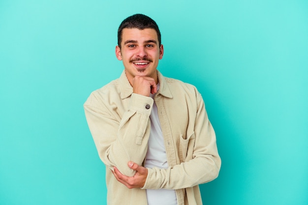 Giovane uomo caucasico isolato sulla parete blu sorridendo felice e fiducioso, toccando il mento con la mano