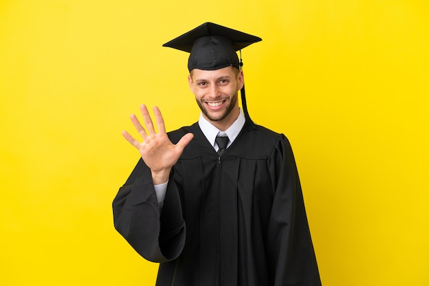 Giovane uomo caucasico laureato isolato su sfondo giallo contando cinque con le dita