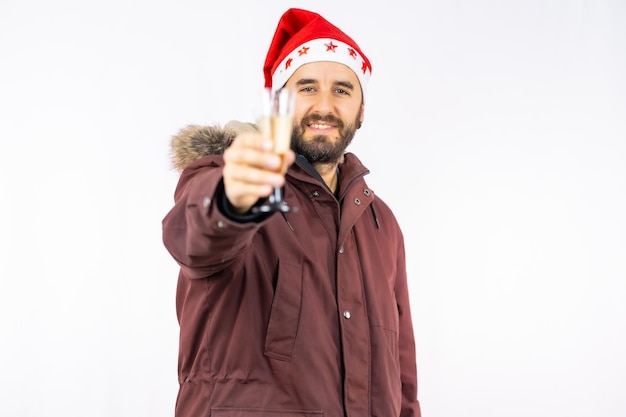 Giovane uomo caucasico molto felice con cappello rosso di Natale che brinda con un bicchiere di champagne su uno sfondo bianco, vestito con una giacca invernale