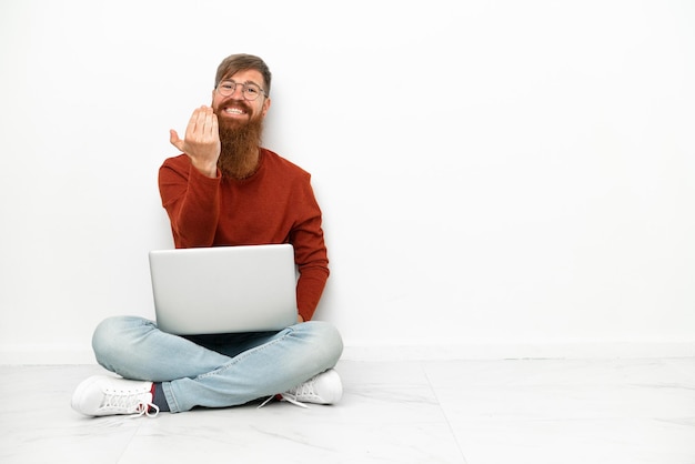 Giovane uomo caucasico rossastro con laptop isolato su sfondo bianco che invita a venire con la mano. Felice che tu sia venuto
