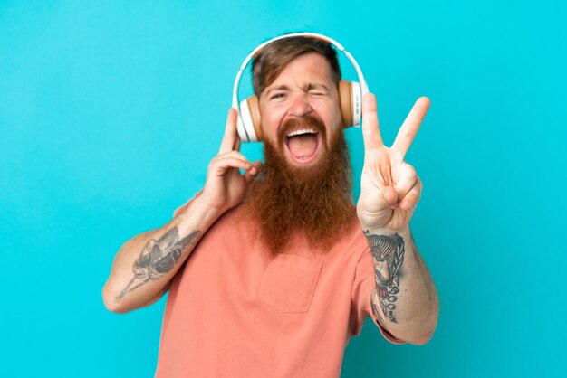 Giovane uomo caucasico rossastro isolato su sfondo blu ascoltando musica e cantando