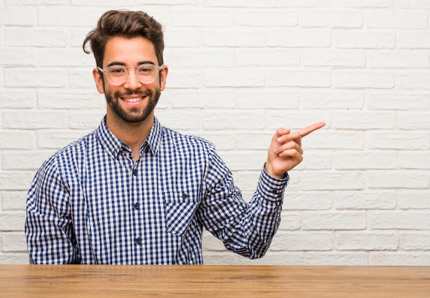 Giovane uomo caucasico seduto che punta verso il lato, sorridente sorpreso presentare qualcosa, naturale e informale