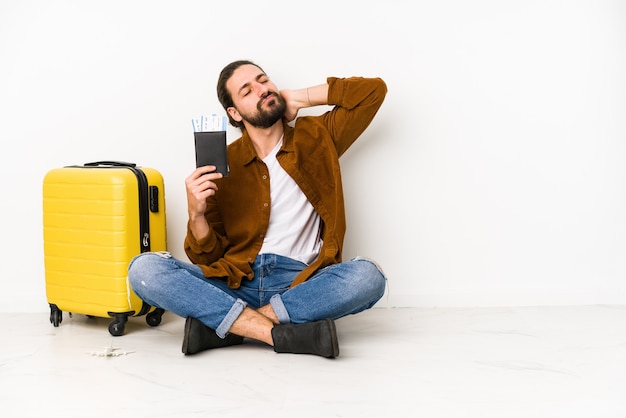 Giovane uomo caucasico seduto in possesso di un passaporto e una valigia isolata toccando la parte posteriore della testa, pensando e facendo una scelta.