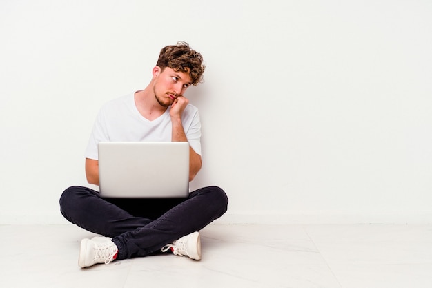 Giovane uomo caucasico seduto sul pavimento che tiene su un laptop isolato su sfondo bianco che si sente triste e pensieroso, guardando lo spazio della copia.