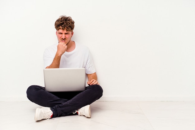 Giovane uomo caucasico seduto sul pavimento che tiene sul computer portatile isolato su sfondo bianco che mostra il pugno alla telecamera, espressione facciale aggressiva.