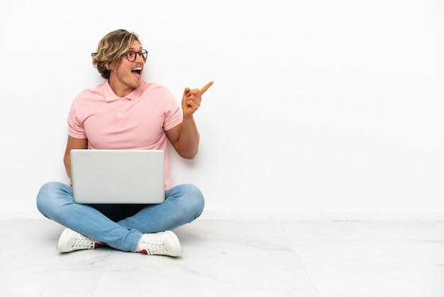 Giovane uomo caucasico seduto sul pavimento con il suo computer portatile isolato su sfondo bianco con l'intenzione di realizzare la soluzione mentre si solleva un dito