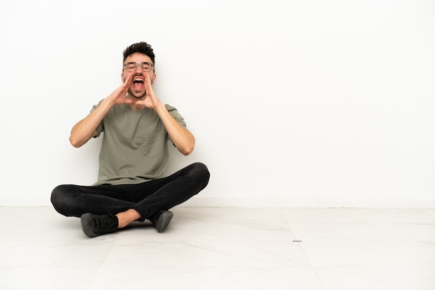 Giovane uomo caucasico seduto sul pavimento isolato su sfondo bianco gridando e annunciando qualcosa