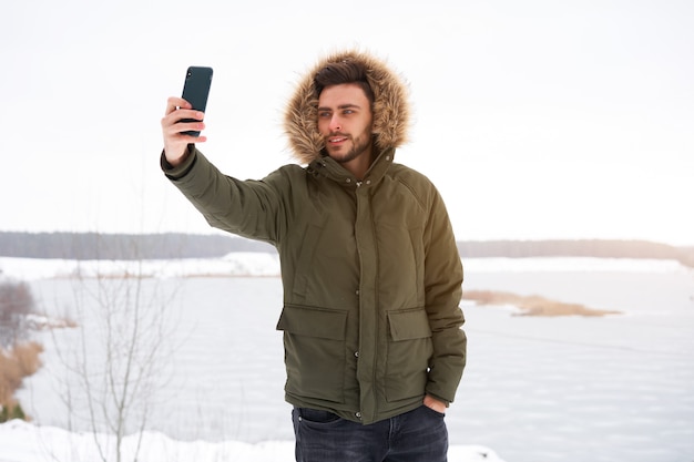 Giovane uomo caucasico sorridente felice in cappuccio della pelliccia Inverno che prende selfie