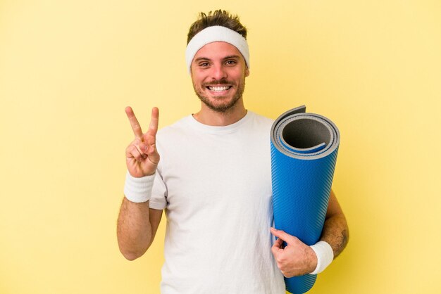 Giovane uomo caucasico sportivo che va a lezioni di yoga mentre tiene un tappetino isolato su sfondo giallo che mostra il numero due con le dita