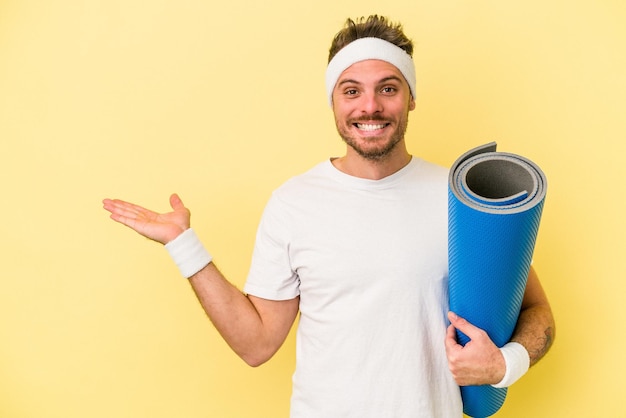 Giovane uomo caucasico sportivo che va a lezioni di yoga mentre tiene un tappetino isolato su sfondo giallo che mostra uno spazio di copia su un palmo e tiene un'altra mano sulla vita.