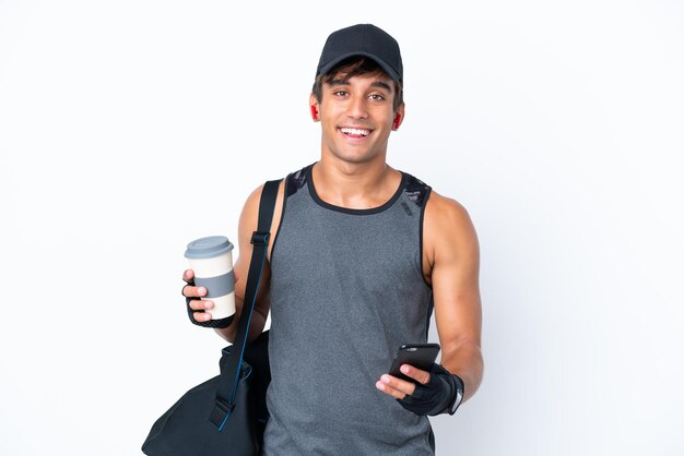 Giovane uomo caucasico sportivo con borsa sportiva isolato su sfondo bianco che tiene caffè da portare via e un cellulare