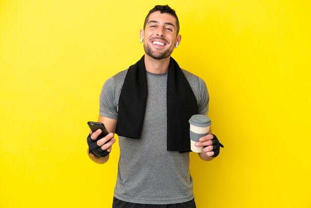 Giovane uomo caucasico sportivo isolato su sfondo giallo che tiene caffè da portare via e un cellulare