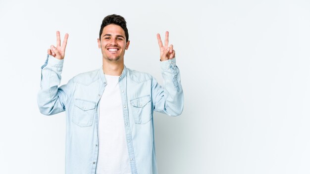 Giovane uomo caucasico su bianco che mostra il segno di vittoria e che sorride ampiamente.