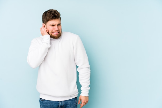 Giovane uomo caucasico sulle orecchie della copertura blu con le mani.