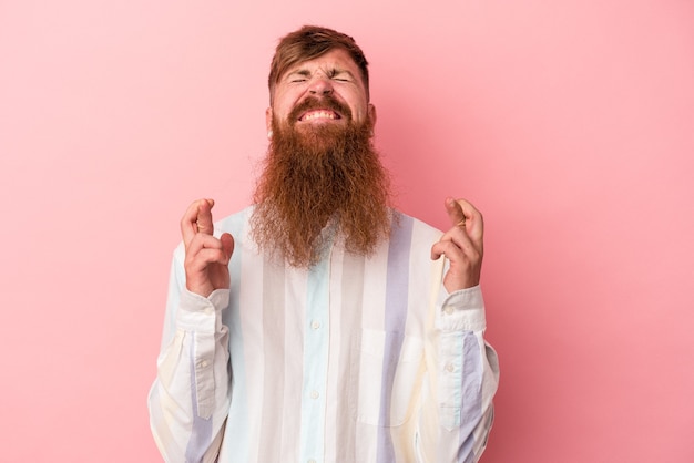 Giovane uomo caucasico zenzero con barba lunga isolato su sfondo rosa incrociando le dita per avere fortuna