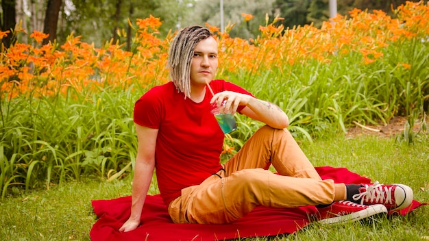 Giovane uomo che beve una limonata rinfrescante seduto sul tappeto nel parco cittadino maschio rilassato con i dreadlocks che beve bibita analcolica attraverso la paglia che disseta riposando sul prato sullo sfondo di fiori sfocati