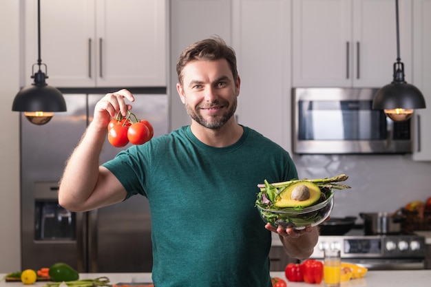 Giovane uomo che cucina insalata sana vegana in cucina uomo millenario al tavolo da cucina moderno tritare vegetab