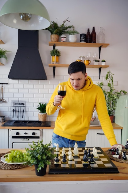 Giovane uomo che gioca a scacchi sul tavolo della cucina e beve vino rosso Maschio con alcol gioca in un gioco da tavolo logico con se stesso in piedi in cucina