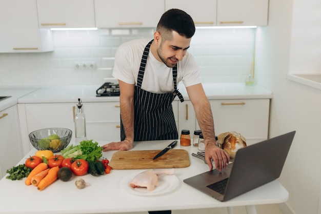 Giovane uomo che guarda la video ricetta sul laptop mentre cucina le verdure in cucina Ritratto di un bel ragazzo in grembiule che impara a cucinare mentre guarda un webinar su un computer nella cucina di casa