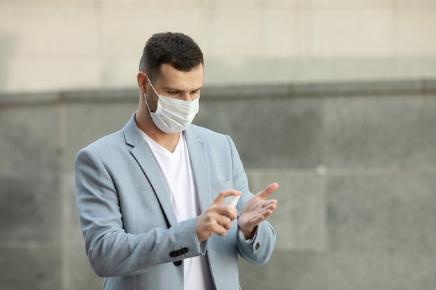 Giovane uomo che indossa la maschera medica utilizzando gel disinfettante per le mani a piedi in città